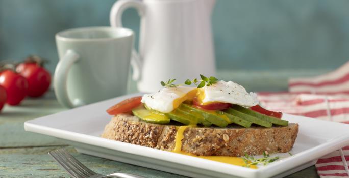 Fotografia em tons de vermelho em uma bancada de madeira verde clara, um paninho vermelho listrado, um prato quadrado branco com uma torrada e um ovo poché em cima com abacate. Ao fundo, tomates, xícara e jarra com café.