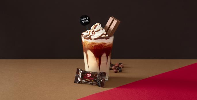 Foto da receita de Frapê Avelã. Observa-se Um fundo escuro marrom e ao centro um copo alto, decorado com calda de chocolate e dentro a bebida cremosa. Ao topo, chantilly e um KIT KAT Dark espetado. Avelãs decoram a parte direita do copo.