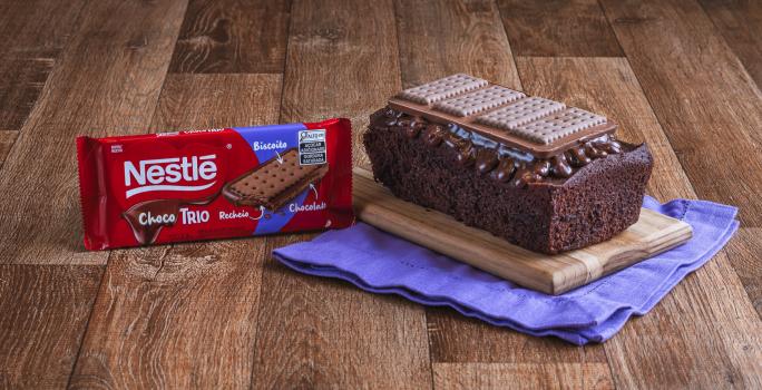 Foto da Receita de Bolo de Chocolate com Cobertura de Choco Trio. Observa-se um bolo em formato inglês sobre uma tábua de madeira e um pano roxo.