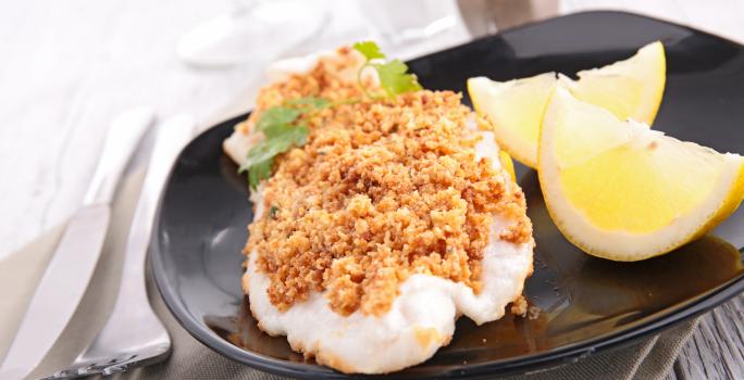Fotografia em tons de marrom em uma bancada com um prato preto oval ao centro e o filé de peixe com uma crosta de aveia e castanhas em cima. Ao lado, pedaços de limão e talheres.