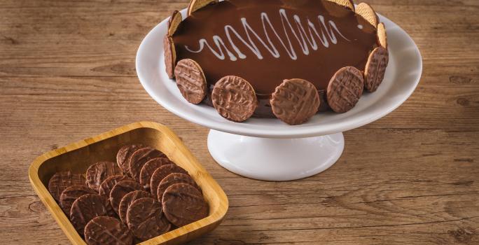 Imagem de uma torta decorada com biscoitos ao redor, disposta num prato branco alto, ao lado de uma tigela de madeira com mais biscoitos dentro