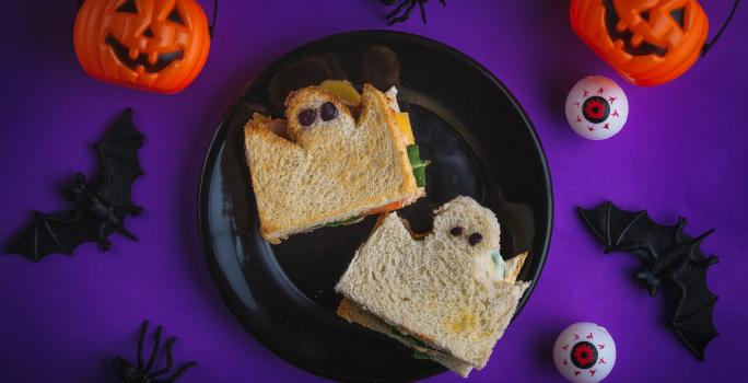 Imagem de um ângulo de cima. Numa mesa em tom roxo escuro há um prato preto com dois sanduíches de pão de forma cortados em formato de fantasmas, com olhos pretos. Ao redor há abóboras, morcegos e aranhas de mentira.