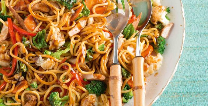 Foto de um prato branco cheio de yakissoba com frango e legumes. Encostado ao prato há duas colheres