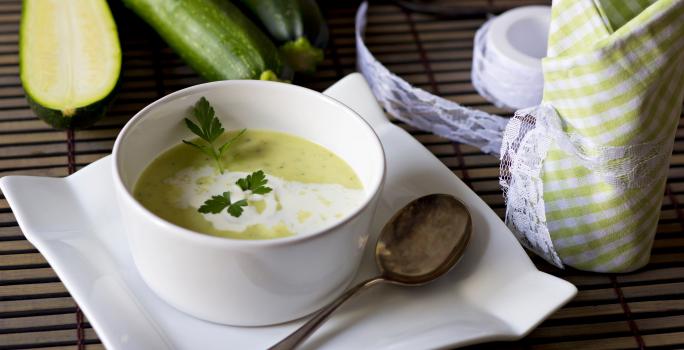 Fotografia de um recipiente fundo branco de vidro com uma sopa de abobrinha e cará, com um creme branco por cima e salsinha. A sopa está em cima de um prato de vidro branco e quadrado, com uma colher de sopa apoiada nele, sobre uma mesa de madeira.