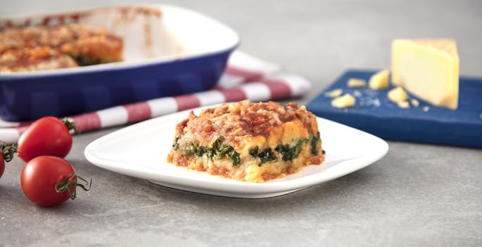 Fotografia em tons de azul em uma bancada de madeira cinza, um pano branco com listras vermelhas, um recipiente azul retangular com a polenta com escarola dentro. Ao centro, um prato branco com uma fatia da polenta com escarola.