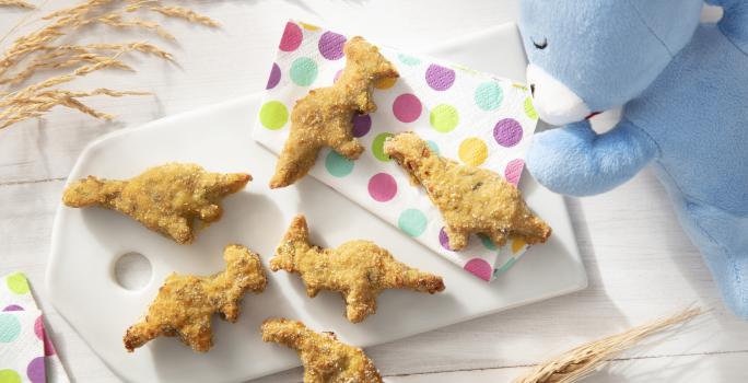 Foto da receita de Nuggets de Frango e Abobrinha. Observa-se uma tábua branca com os nuggets em formato de dinossauro sobre ela em cima de guardanapos coloridos de bolinha.