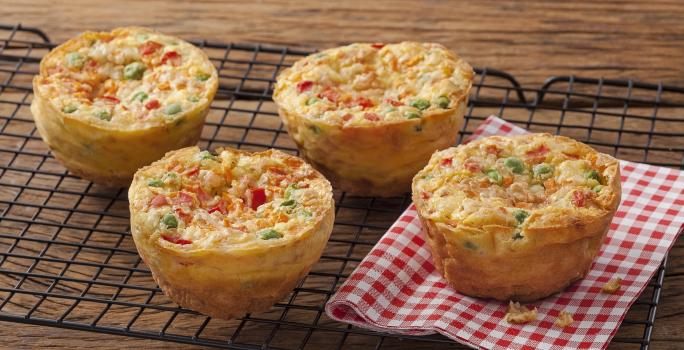 Fotografia em tons de vermelho em uma bancada de madeira com um pano xadrez vermelho com quatro muffins de tapioca com legumes.