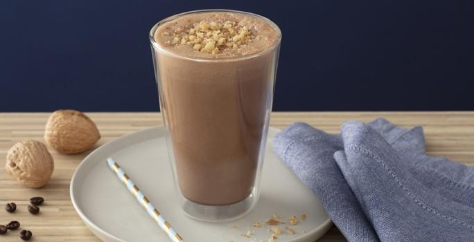 Foto em tons de marrom da receita de smoothie de café com nozes servida em um copo alto sobre uma base de porcelana cinza com um canudo listrado ao lado. Como decoração há duas nozes e um paninho azul