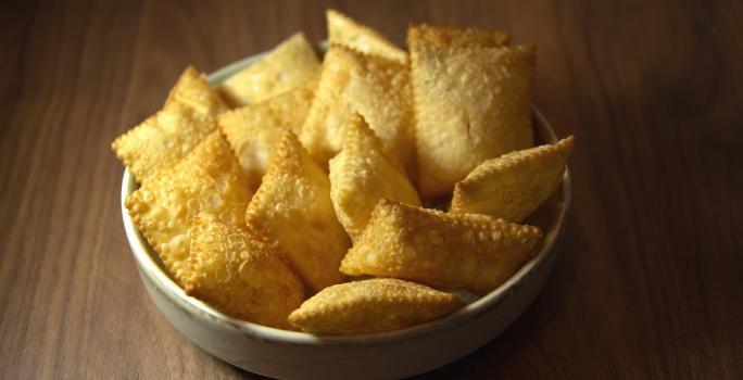 Fotografia em tons de amarelo, em uma bancada de madeira de cor marrom. Ao centro, um recipiente branco contendo os pasteis.