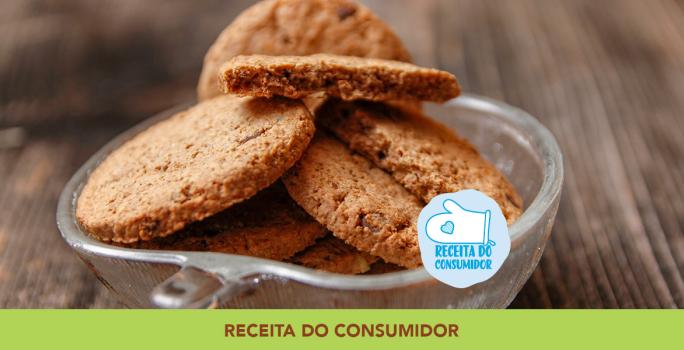 Foto aproximada da receita de Cookies Integrais, dentro de um pote de vidro, em tom marrom claro, sobre uma bancada de madeira e com dizeres no rodapé em tom verde.