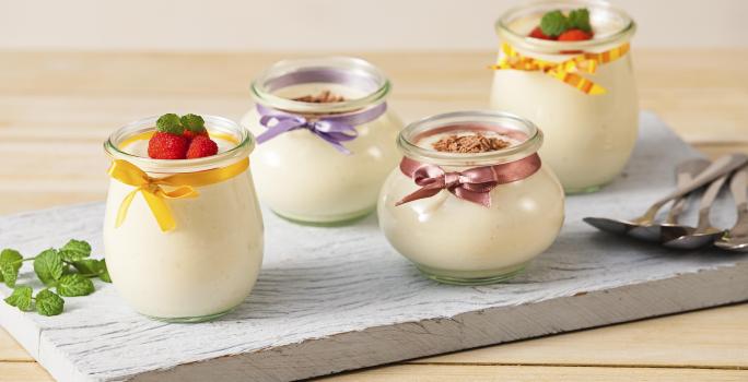 Fotografia em tons de branco e cinza, ao centro  quatro potinhos sobre tábua cinza, decorados com fitas coloridas, frutas, hortelã e raspas de chocolate e contendo mousse, ao lado ramo de hortelã e quatro colheres, tudo sobre bancada de madeira.