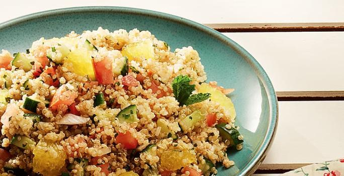 Fotografia em tons de verde e amarelo, com porcelana verde contendo tabule de quinoa com legumes e gomos de laranja, decorado com folhas de hortelã, tudo sobre bancada de madeira bege.