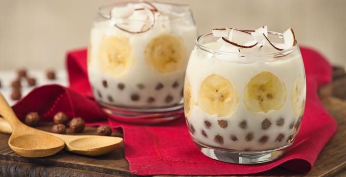 Fotografia em tons de vermelho e marrom em uma bancada branca com uma tábua de madeira escura, um pano vermelho, duas taças de vidro com o docinho de banana com Nescau Cereal.