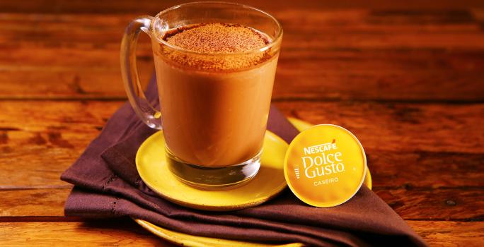 Fotografia em tons de amarelo em uma bancada de madeira escura, um prato amarelo redondo, um pano marrom e uma xícara de vidro com o chocafé com chocolate dentro dela.