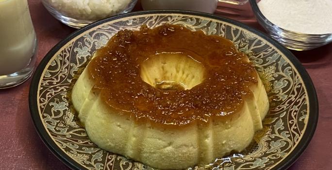 Foto em tons de bege escuro da receita de pudim de arroz inteira servida sobre um prato grande decorado. Ao fundo há alguns recipientes com arroz, açúcar, leite ninho e leite MOÇA.