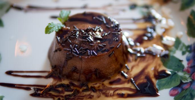 Fotografia de um mini pudim de chocolate sem furo no meio, decorado com granulado, calda de chocolate e uma folha de hortelã, sobre um prato branco desenhado..