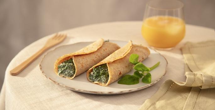 Foto em tons de bege da receita de panqueca de espinafre com ricota servida em duas porções sobre um prato de cerâmica claro em cima de uma mesa decorada com uma toalha bege, um copo de vidro com suco de laranja, um paninho verde e um garfo de madeira