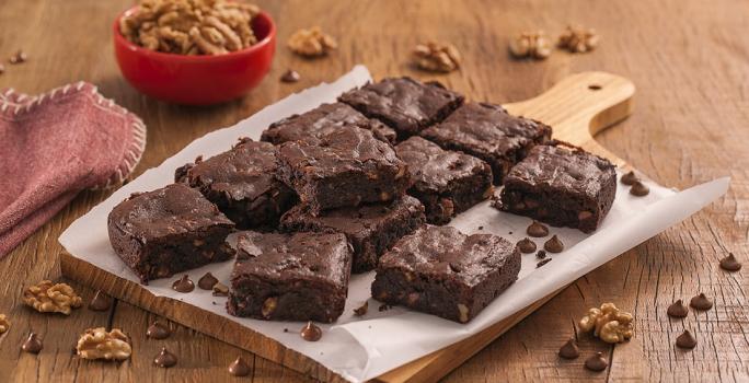 Fotografia em tons de marrom e vermelho de uma bancada de madeira com uma tábua e um papel manteiga, sobre ele pedaços de brownie. Ao fundo um recipiente vermelho pequeno com nozes e um pacote de brownie dois frades.