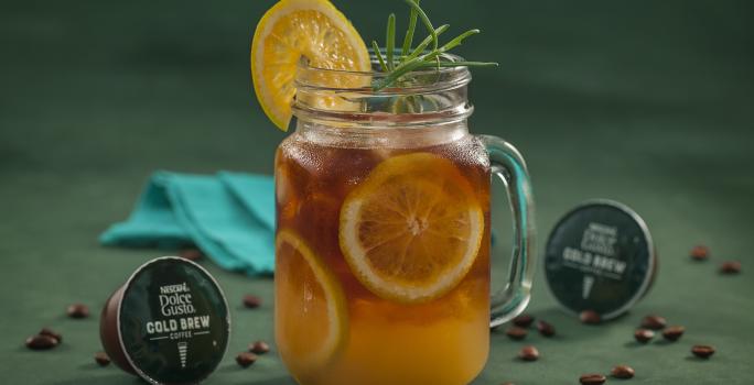 Fotografia em tons de verde de uma bancada verde, sobre ela um copo de vidro com cold brew com laranjas. Ao lado capsulas de Dolce Gusto Cold Brew e um paninho azul.
