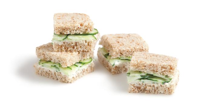 Fotografia em tons de verde em um fundo branco com quatro mini sanduichinhos no pão integral com recheio de pepino.