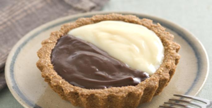 Fotografia em tons de bege em uma mesa de madeira com um pano bege xadrez, um prato redondo raso bege com detalhe azul e a tortinha de leite condensado Moça e chocolate em cima dele.