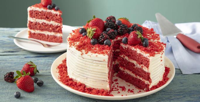 Fotografia em tons de vermelho em uma bancada de madeira clara, um prato redondo branco com o bolo red velvet cortado ao meio. Ao fundo, um prato pequeno com o pedaço.