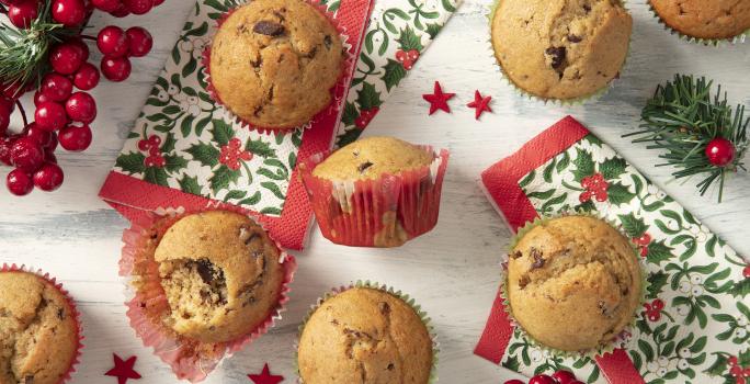 Foto da Receita de Muffin Natalino feito na Air Fryer. Observa-se 7 muffins de baunilha com chocolate em forminhas natalinas vistos de cima. Decorações natalinas enfeitam a foto, como guardanapos e decorações de árvore de Natal.