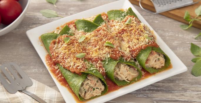 Foto da receita de Panqueca Verde com Frango Cremoso. Observa-se um refratário branco com três panquecas verdes, com molho vermelho por cima, queijo ralado e manjericão. Tomates, manjericão, um mini ralador e queijo ralado decoram a foto atrás.