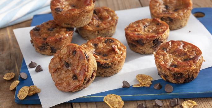 Fotografia em tons de azul em uma bancada de madeira com uma tábua azul, com um folha de papel manteiga e vários muffins de banana com gotas de chocolate em cima dela. Nesfit Cereais Matinais e algumas gotas de chocolate espalhadas pela bancada.