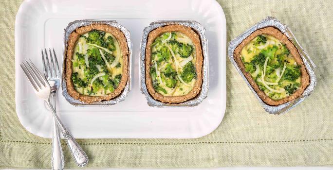 Fotografia em tons de branco, verde e dourado de uma bancada vista de cima mostrando refratário branco com duas porções de quiche com brócolis e dois garfos, com mais uma porção ao lado, tudo sobre guardanapo em tom de branco.