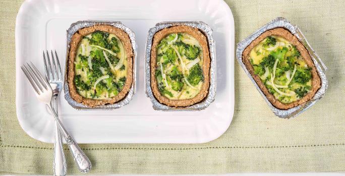 Fotografia em tons de branco, verde e dourado de uma bancada vista de cima mostrando refratário branco com duas porções de quiche com brócolis e dois garfos, com mais uma porção ao lado, tudo sobre guardanapo em tom de branco.