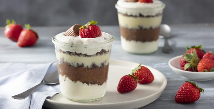 Fotografia em tons de vermelho em uma bancada de madeira clara, um paninho bege, um copo de vidro com a sobremesa de creme branco e chocolate dentro dele e em cima de um prato branco redondo. Ao fundo, mais um copo e morangos para decorar.