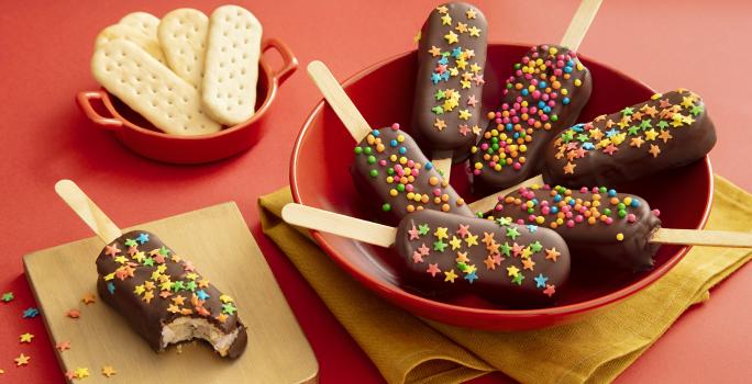 Foto da receita de Pirulito de Biscoito. Observa-se 7 pirulitos decorados com chocolate e confeitos em um prato fundo vermelho sobre um guardanapo de pano amarelo