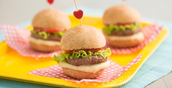 3 mini hambúrguer por cima de uma bandeja amarela. Abaixo um pano azul claro. Fotografia tirada de frente de uma bancada cinza.