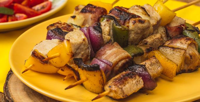 Foto bem aproximada de um prato amarelo sobre o qual há quatro espetinhos montados com carne de porco, cebolas roxa e tradicional e pimentões verdes e amarelos. Ao fundo há um prato branco com uma salada de tomates.