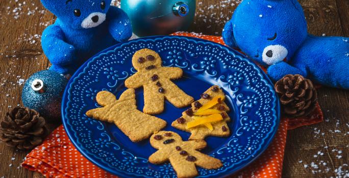 Fotografia em tons de laranja e azul em uma bancada de madeira, um paninho laranja, um prato azul com os biscoitinhos natalinos em cima dele. Ao lado, decoração e enfeites de natal.