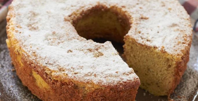 em uma mesa de madeira contém um pano xadrez nos tons vinho e cinza e por cima do pano o bolo de fubá coberto com açúcar de confeiteiro.