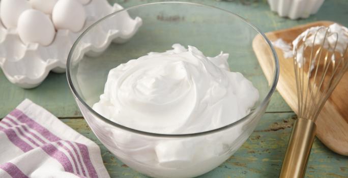 Fotografia em tons de verde em uma bancada de madeira verde, um pano branco com listras roxas, um recipiente de vidro com o merengue dentro dele. Ao fundo, potinhos com docinhos cobertos com merengue, ovos, tábua de madeira com um fouet.