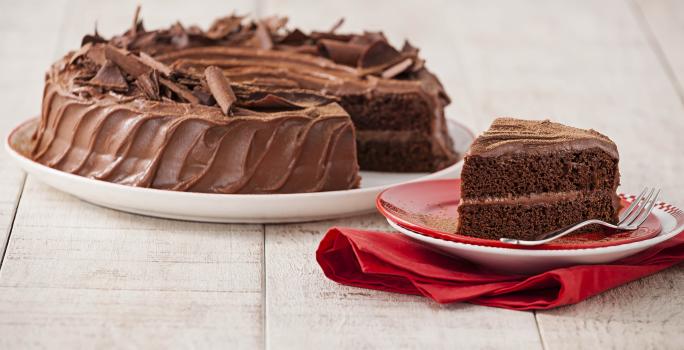 Fotografia em tons chocolate e branco, ao centro prato com bolo de chocolate fatiado, fatia ao lado sobre pratos vermelho e branco com um garfo e sobre guardanapo vermelho, tudo sobre bancada de madeira em pátina.