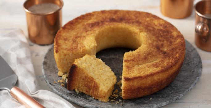 Fotografia em tons de dourado em uma bancada com uma toalha branca com detalhes cinzas, um prato redondo grande de pedra cinza e o bolo de queijo com coco em cima dele e uma fatia cortada. Ao lado, uma espátula com cabo cobre e ao fundo, várias xícaras.