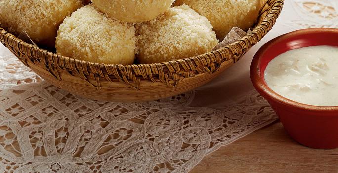 Fotografia em tons de branco, marrom, pardo e vermelho de uma bancada vista de cima. Um pano de renda branco e por cima um recipiente cor pardo e redondo com pães bolinha, ao lado um potinho vermelho redondo com creme branco.