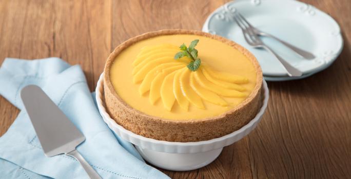 fotografia em tons de marrom, branco e amarelo de uma bancada marrom vista de frente, contém uma bailarina para servir sobremesa branca com uma torta por cima. Ao lado esquerdo contém um prato redondo branco e do lado direito um pano azul com uma espátula