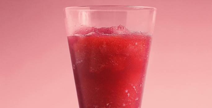 Fotografia em tons de vermelho e rosa de uma bancada rosa sobre ela um copo de vidro com suco de melancia.