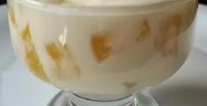 Foto da receita de mousse de abacaxi servida em uma taça de vidro com pedaços de abacaxi à mostra sobre um prato de porcelana branco