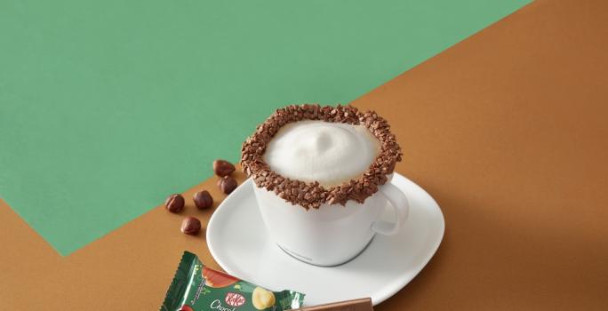 Foto da receita de cappuccino kit kat servida em uma xícara branca com um kit kat avelã ao lado em fundo marrom e verde