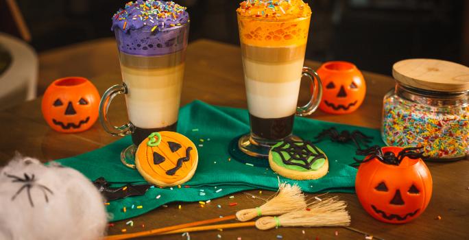 Fotografia em tons de laranja de uma mesa de madeira, duas bebidas, uma decorada com chantilly roxo e uma decorada com chantilly laranja. Em volta biscoitos decorados de abóbora, teia de aranha e elementos de halloween.