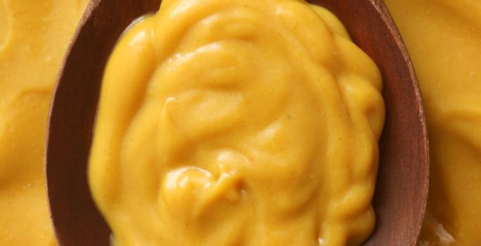 Fotografia vista de cima de um molho de mostarda em uma colher de madeira escura, apoiada sobre mais molho de mostarda.