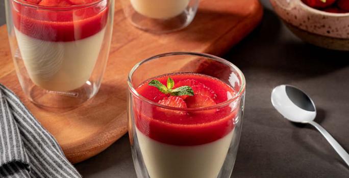 Imagem de três copos pequenos contendo a receita de Panacota com calda de morango. Dois estão sobre uma tábua com um tecido ao lado e um está mais à frente. Ao fundo há um pote com morangos e uma colher de metal