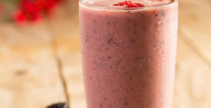 Fotografia em tons de rosa em uma mesa de madeira com um copo de vidro ao centro com o shake de frutas vermelhas dentro. Morangos e mirtilos espalhados pela mesa.