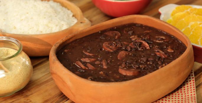 Fotografia em tons de bege em uma bancada de madeira com um pano branco com bolinhas laranjas, um recipiente de barro com a feijoada dentro. Ao lado, potes de barro com arroz, farofa e laranja dentro de cada um.